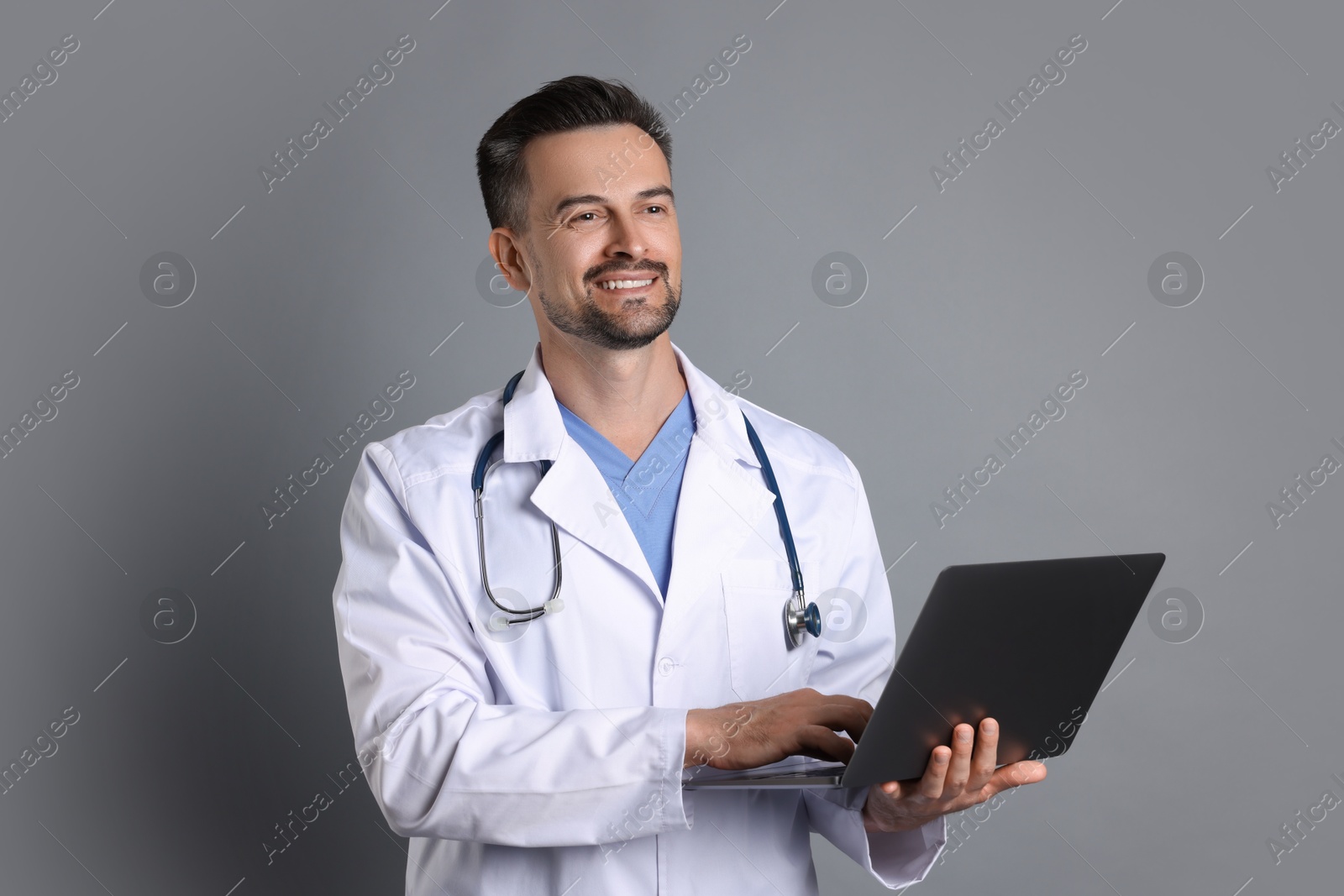 Photo of Smiling doctor with stethoscope and laptop on grey background