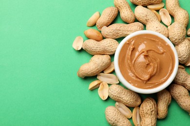 Tasty peanut butter in bowl and groundnuts on green background, flat lay. Space for text