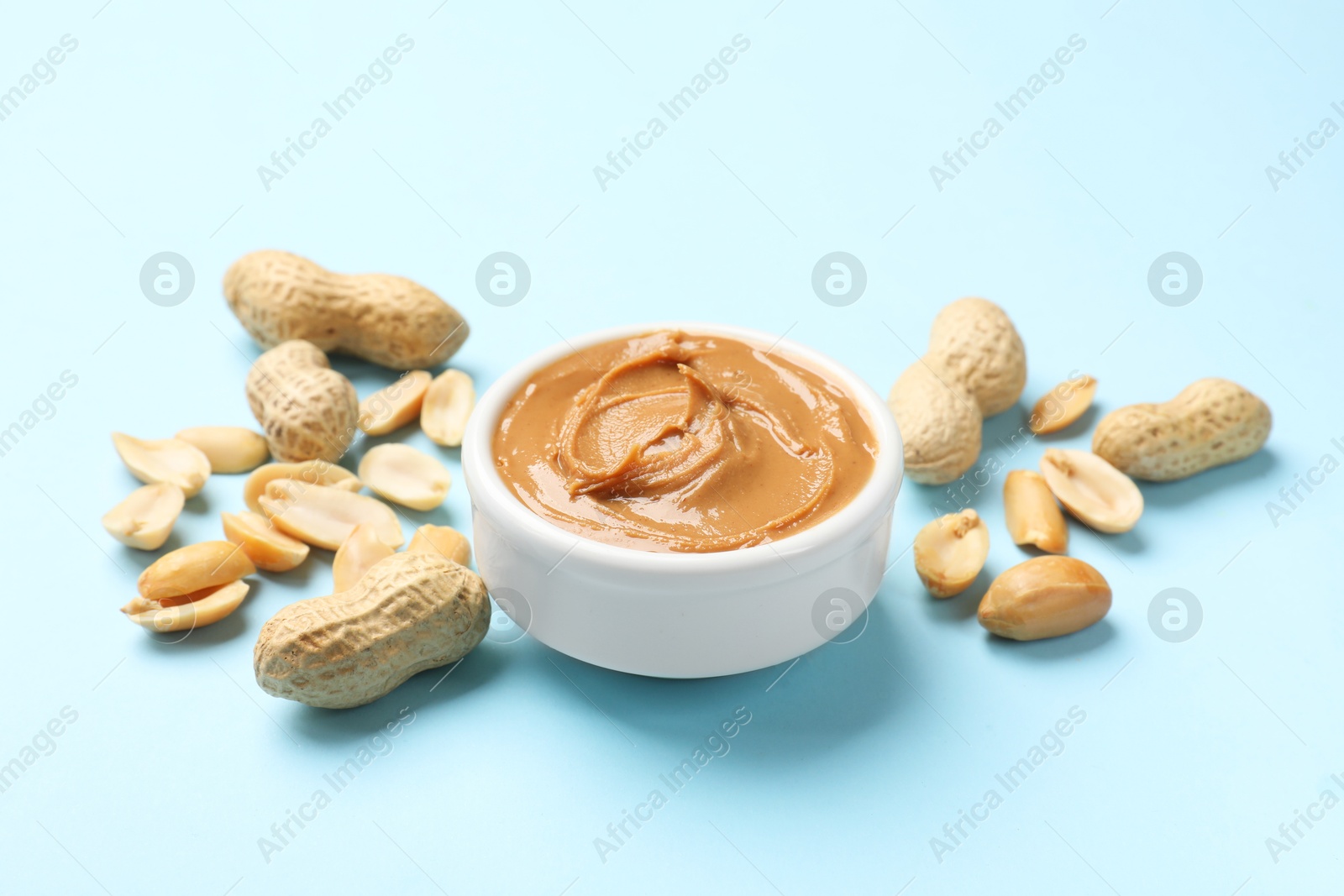 Photo of Tasty peanut butter in bowl and groundnuts on light blue background