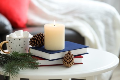 Photo of Burning candle, books, fir branches and cones on coffee table indoors. Christmas atmosphere