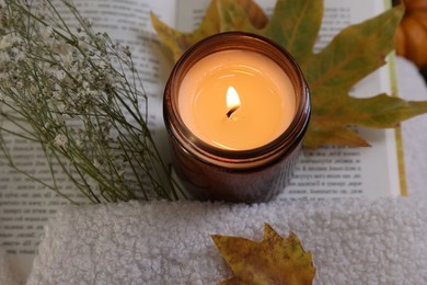 Photo of Burning candle, open book, sweater and autumn decor on table, closeup