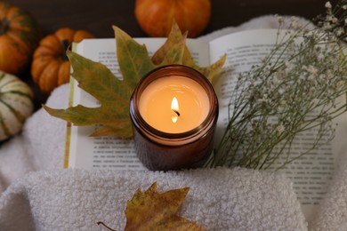 Photo of Burning candle, open book, sweater and autumn decor on table, closeup