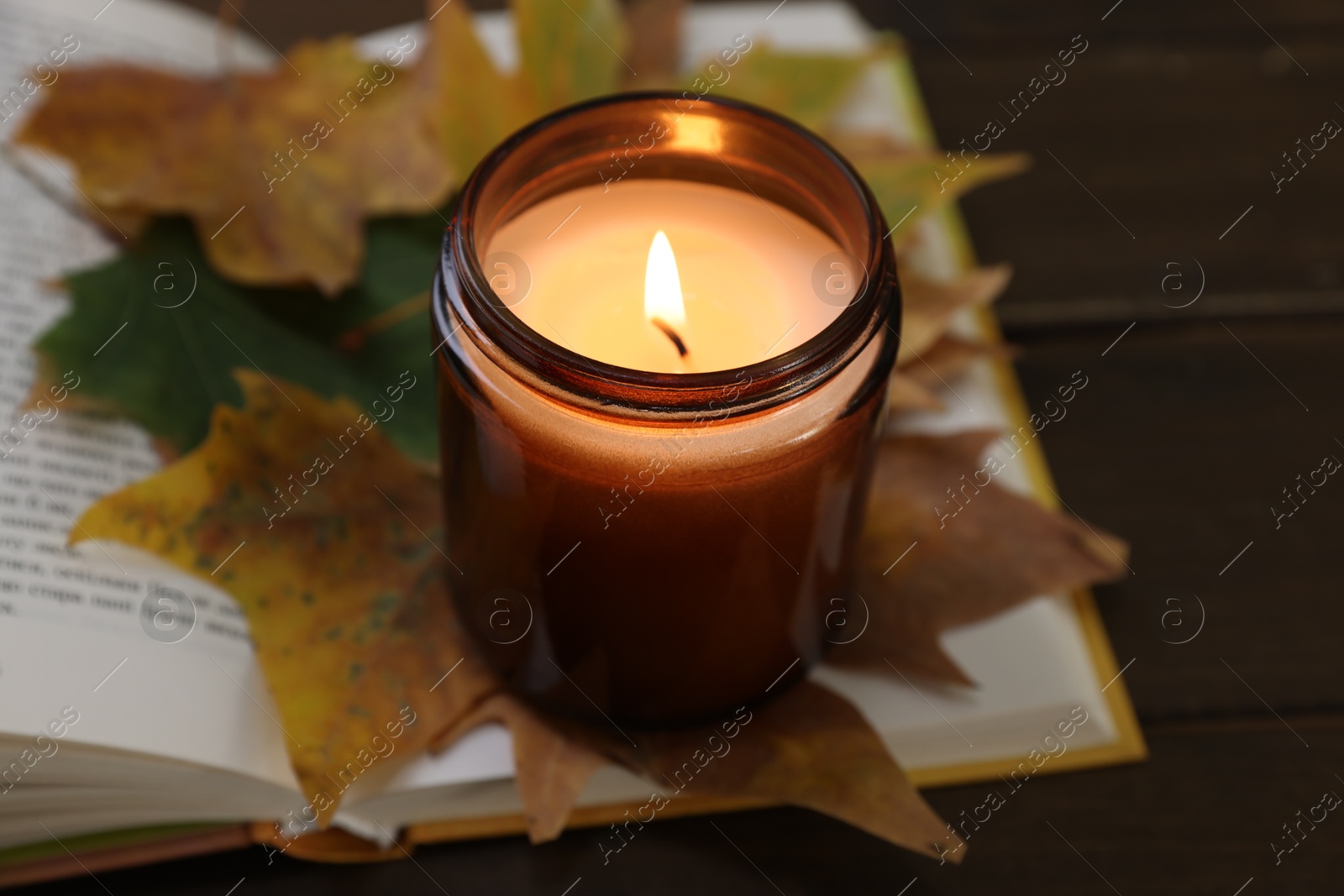 Photo of Burning candle, dry leaves and open book on wooden table, closeup. Autumn atmosphere