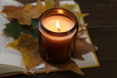 Photo of Burning candle, dry leaves and open book on wooden table, closeup. Autumn atmosphere