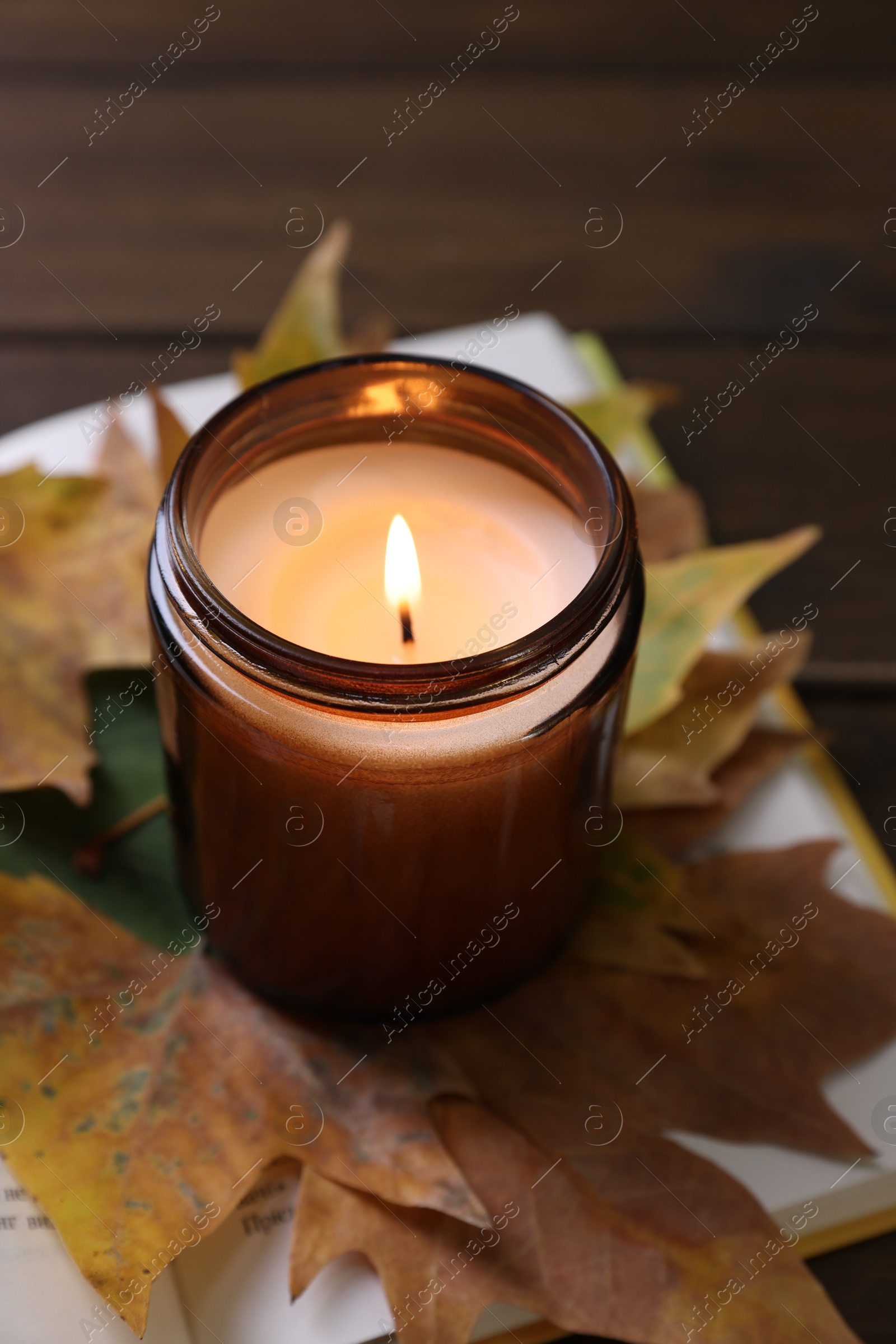 Photo of Burning candle, dry leaves and open book on wooden table, closeup. Autumn atmosphere