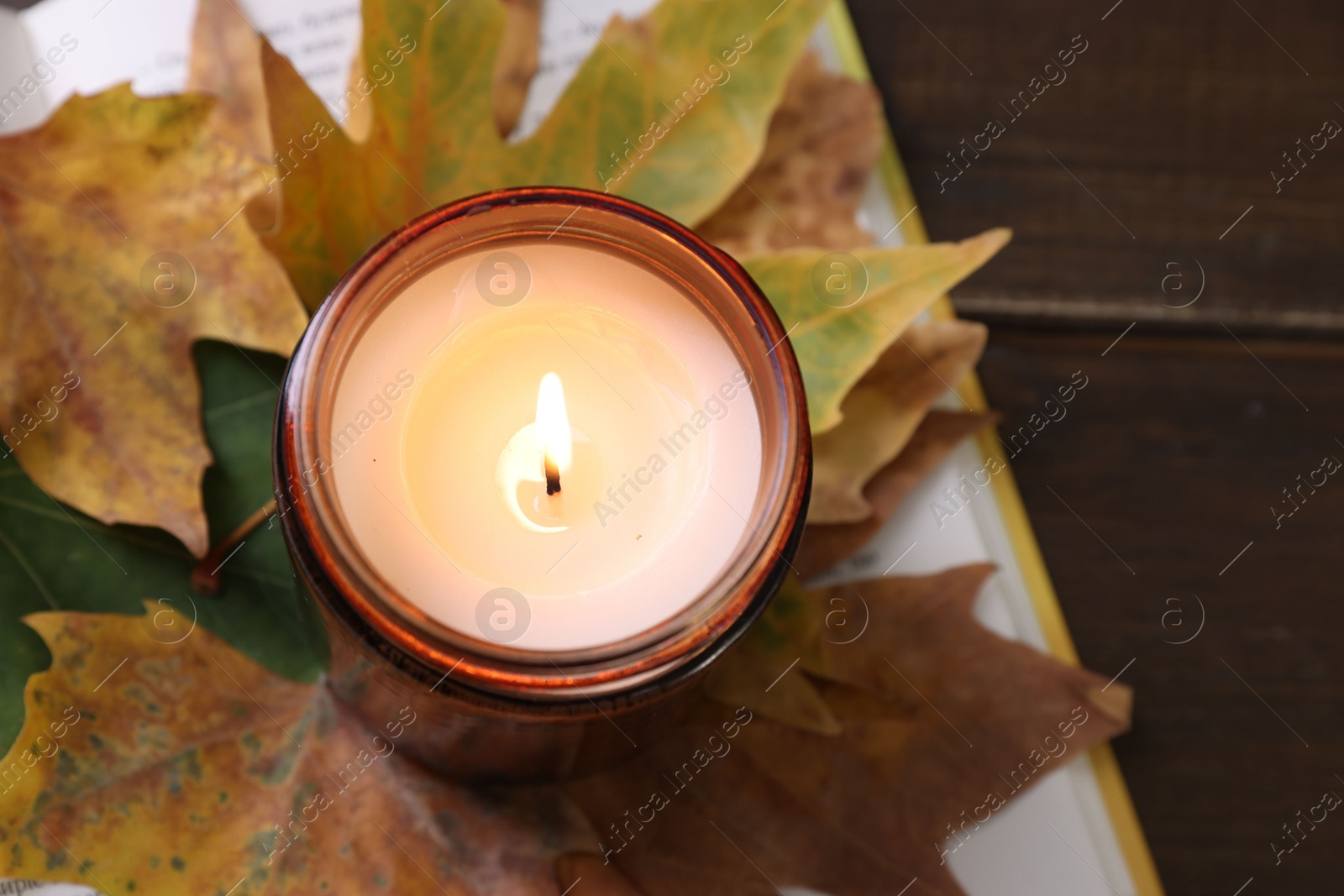 Photo of Burning candle, dry leaves and open book on wooden table, closeup. Autumn atmosphere