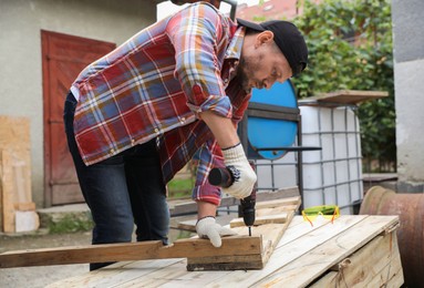 Man twisting screw into wooden plank with electric screwdriver outdoors