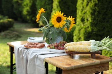 Vase with sunflowers, grapes, pie and corncobs on wooden table in garden