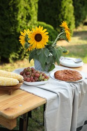 Vase with sunflowers, grapes, pie and corncobs on wooden table in garden