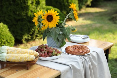 Photo of Vase with sunflowers, grapes, pie and corncobs on wooden table in garden