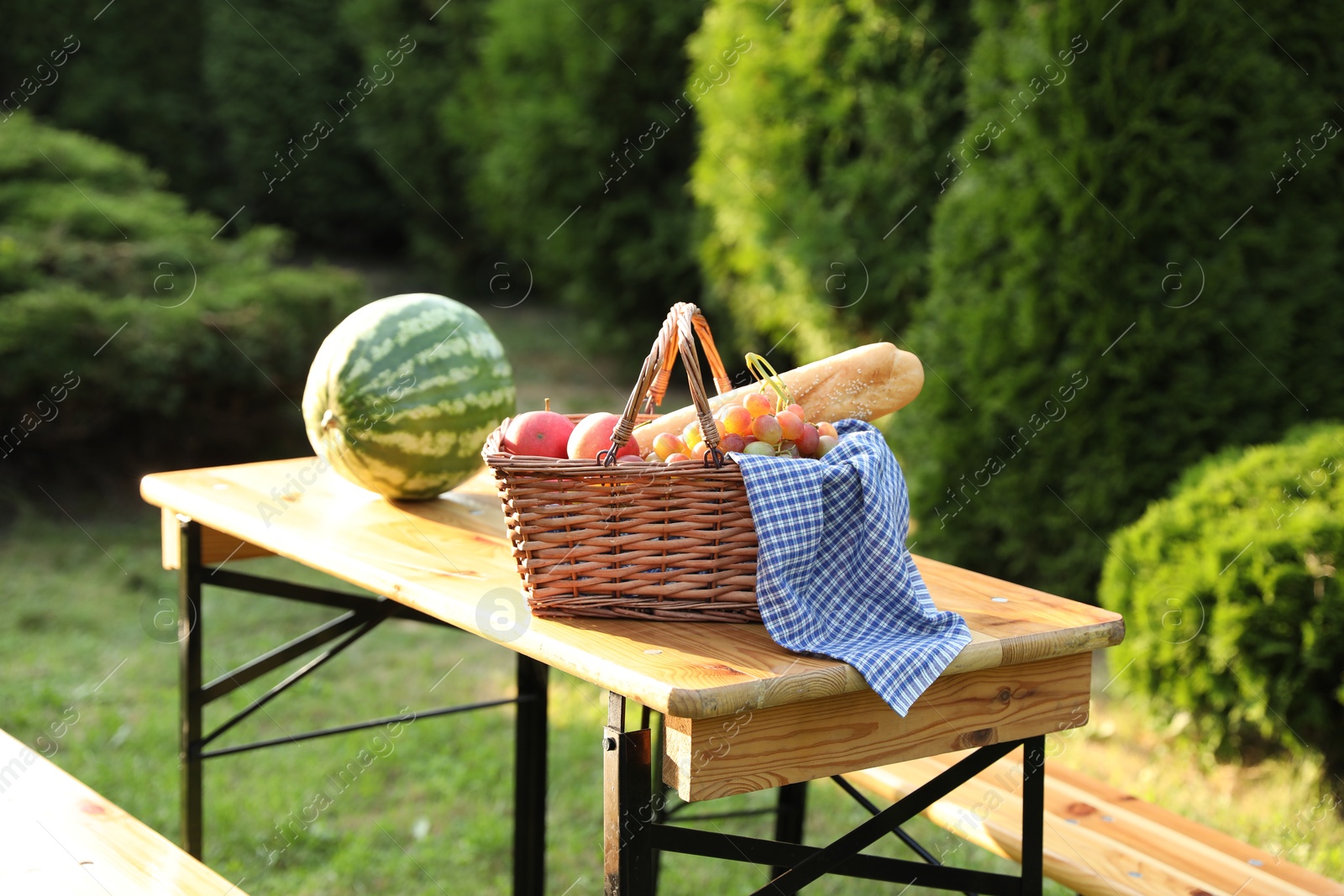 Photo of Wicker basket with grapes, apples and baguette on wooden table in garden