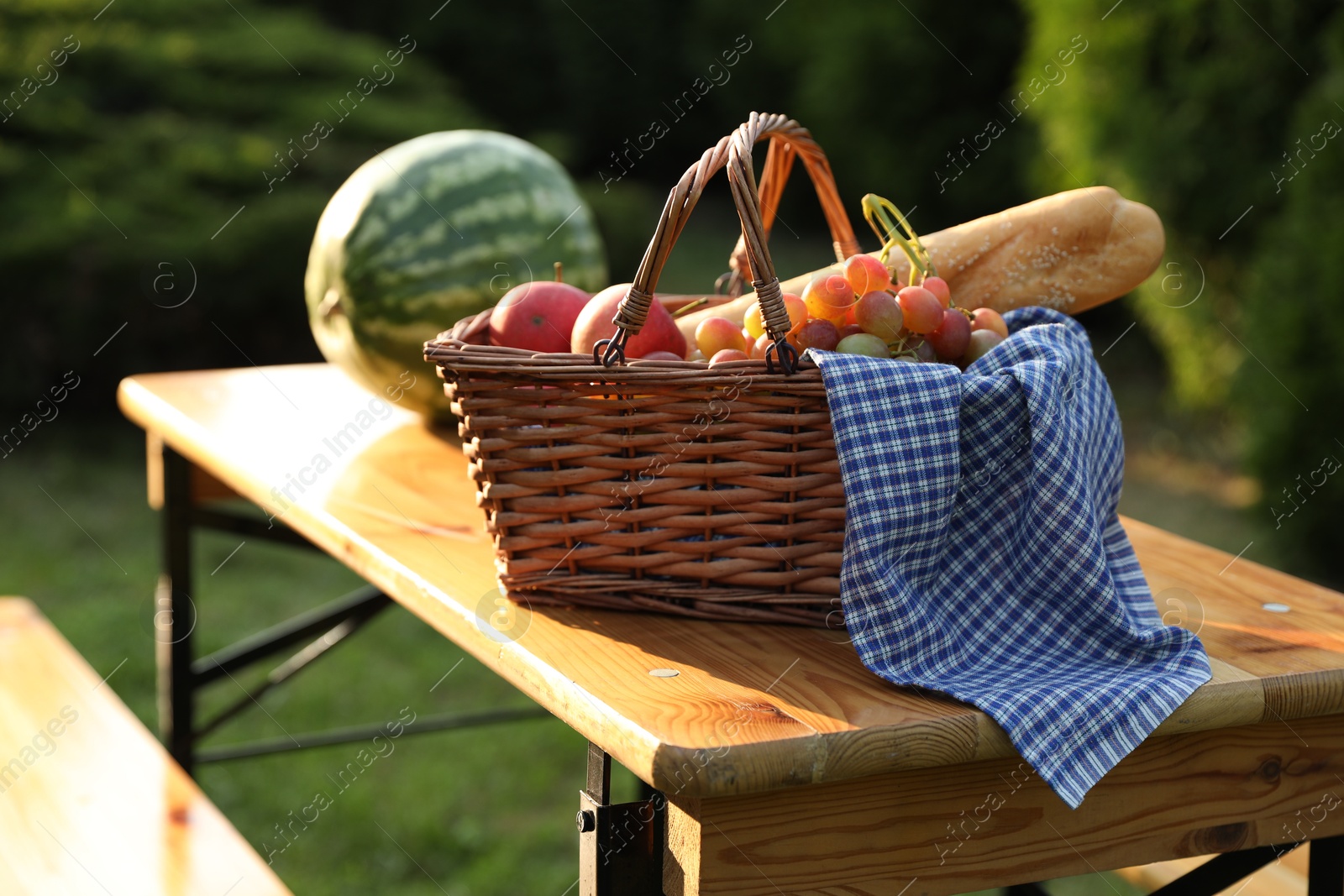 Photo of Wicker basket with grapes, apples, baguette and watermelon on wooden table in garden