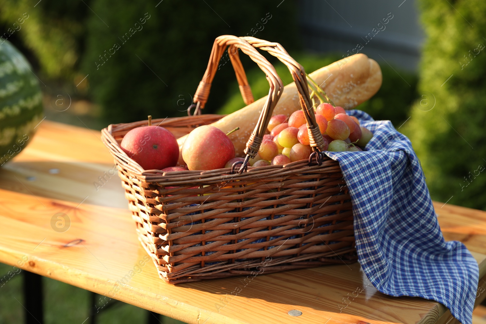 Photo of Wicker basket with grapes, apples and baguette on wooden table in garden