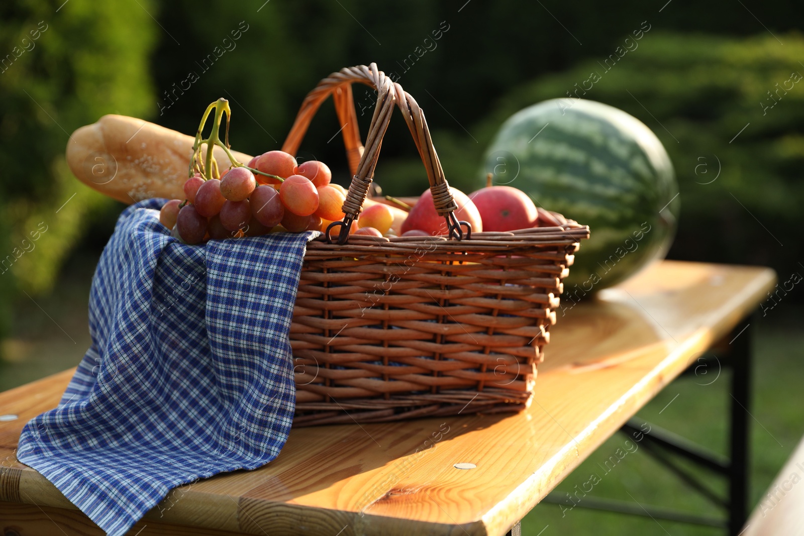 Photo of Wicker basket with grapes, apples, baguette and watermelon on wooden table in garden