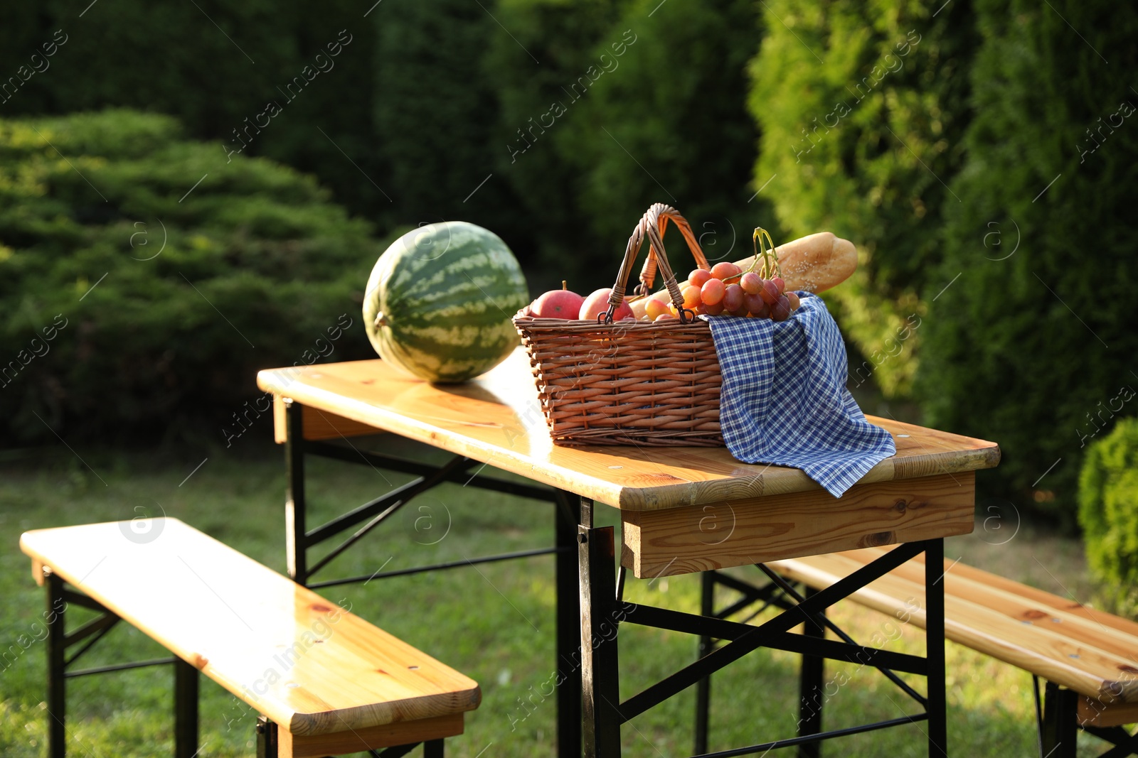 Photo of Wicker basket with grapes, apples, baguette and watermelon on wooden table in garden