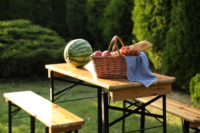 Wicker basket with grapes, apples, baguette and watermelon on wooden table in garden