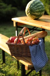 Photo of Wicker basket with grapes, apples and baguette on wooden bench near table in garden