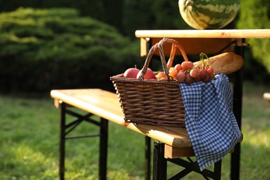 Photo of Wicker basket with grapes, apples and baguette on wooden bench near table in garden