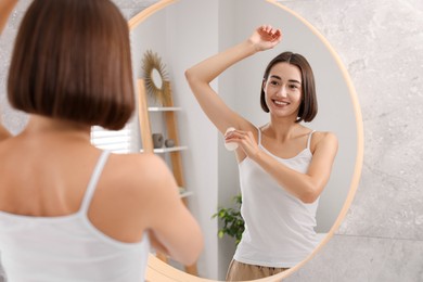 Photo of Smiling woman applying roll-on deodorant near mirror at home, back view