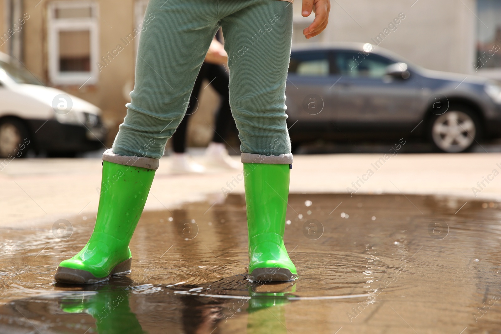 Photo of Little girl wearing green rubber boots standing in puddle outdoors, closeup