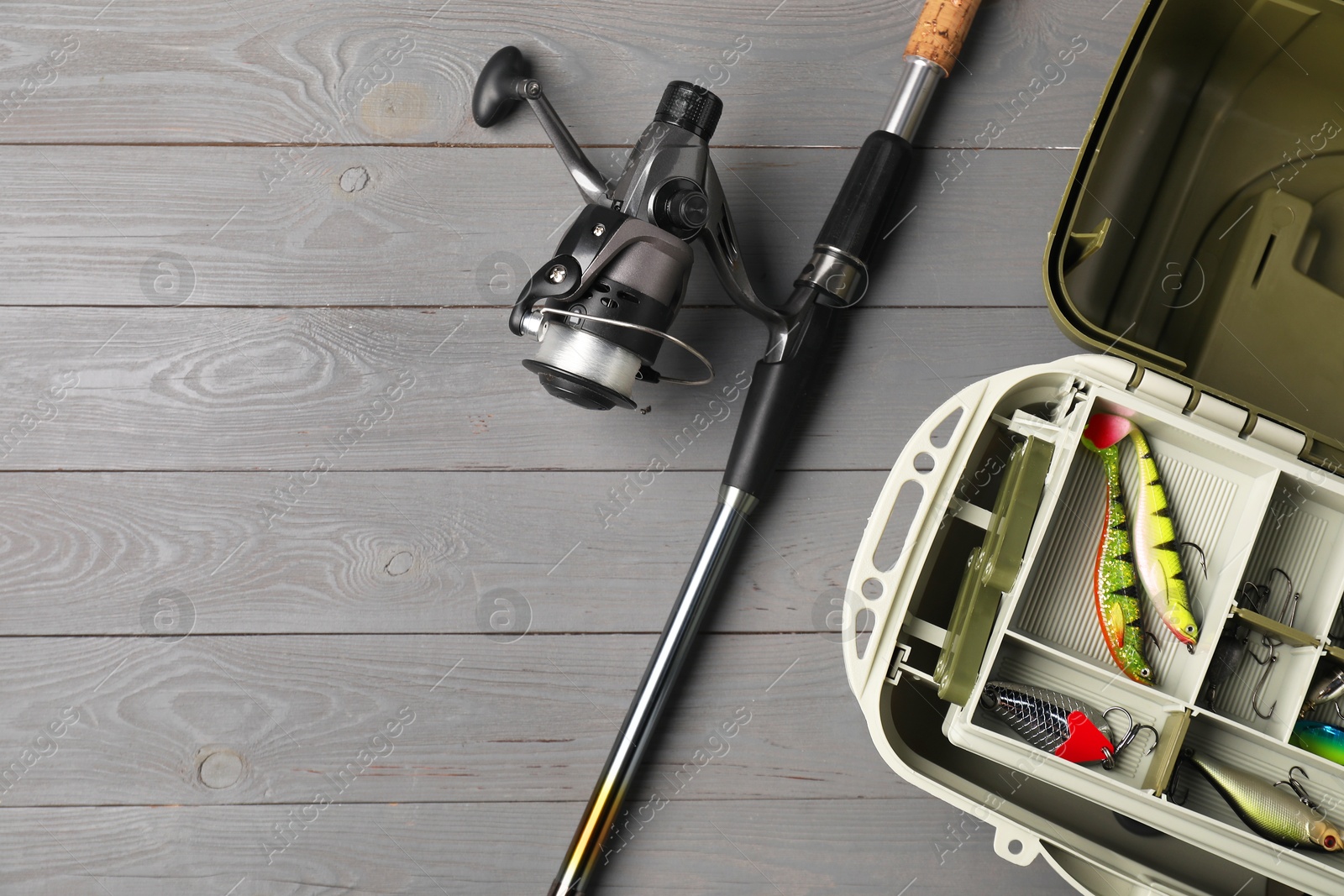 Photo of Different fishing tackles in box and rod with spinning reel on grey wooden table, top view. Space for text