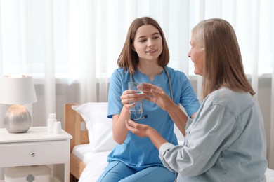 Photo of Young healthcare worker giving glass of water to senior patient on bed indoors