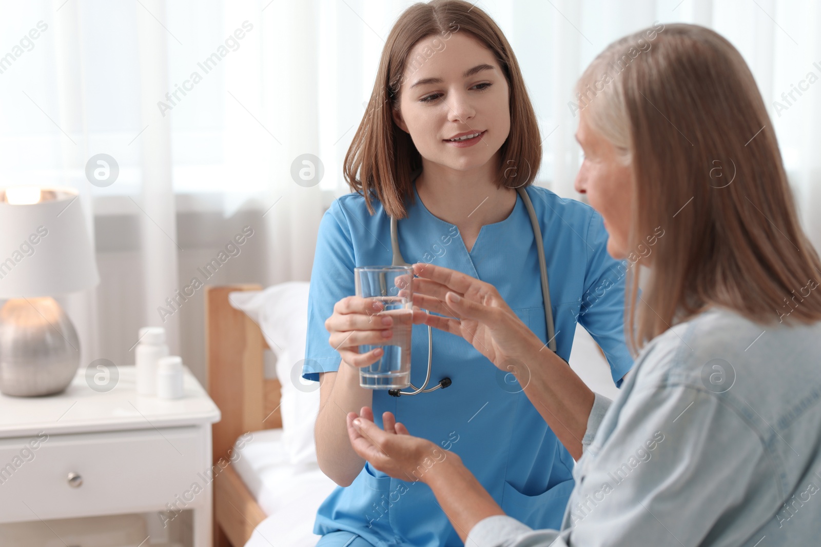 Photo of Young healthcare worker giving glass of water to senior patient on bed indoors