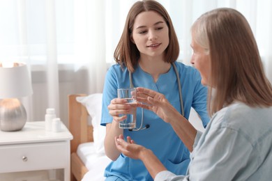 Photo of Young healthcare worker giving glass of water to senior patient on bed indoors