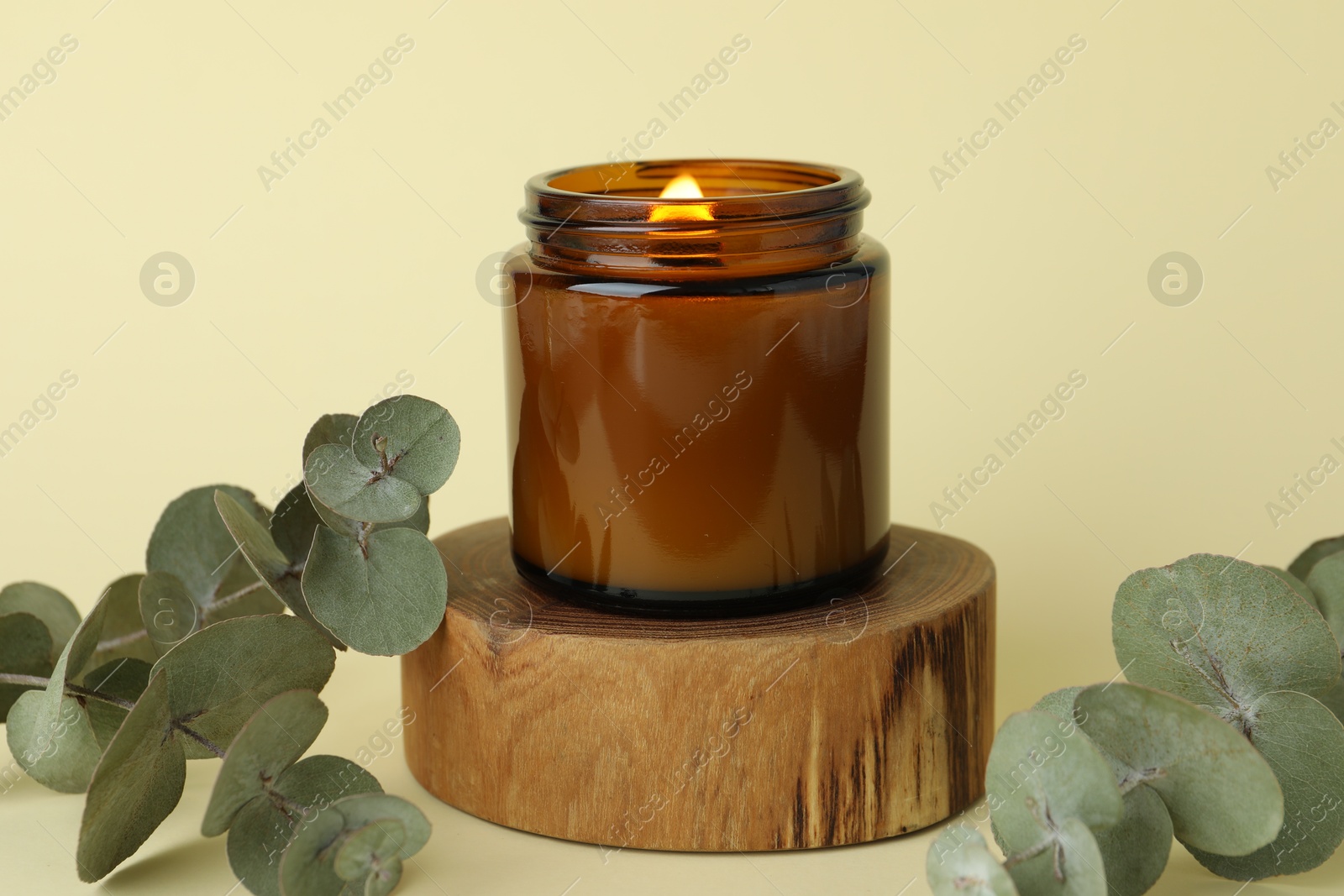 Photo of Beautiful burning candle and eucalyptus branches on pale yellow background, closeup