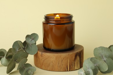 Beautiful burning candle and eucalyptus branches on pale yellow background, closeup