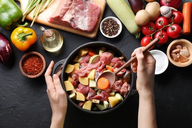 Photo of Cooking stew. Woman with uncooked meat, vegetables and pot on black table, top view