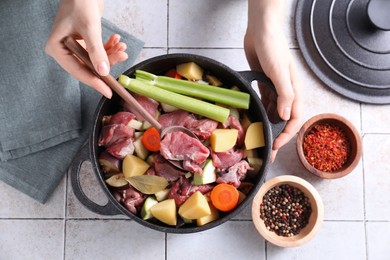 Photo of Woman preparing stew with vegetables and meat at white tiled table, top view