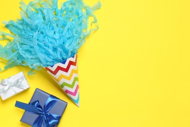 Photo of Party hat with beautiful tissue paper and gift boxes on yellow background, flat lay. Space for text