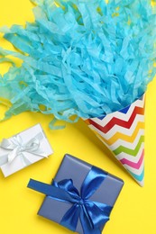 Party hat with beautiful tissue paper and gift boxes on yellow background, flat lay