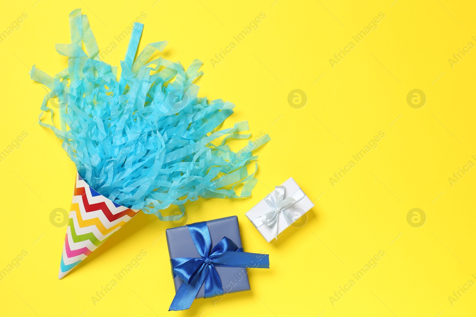 Photo of Party hat with beautiful tissue paper and gift boxes on yellow background, flat lay. Space for text