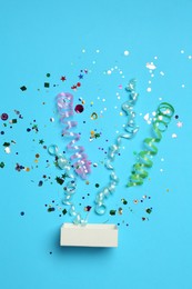 Photo of Shiny confetti and party streamers flying out of little box on light blue background, flat lay