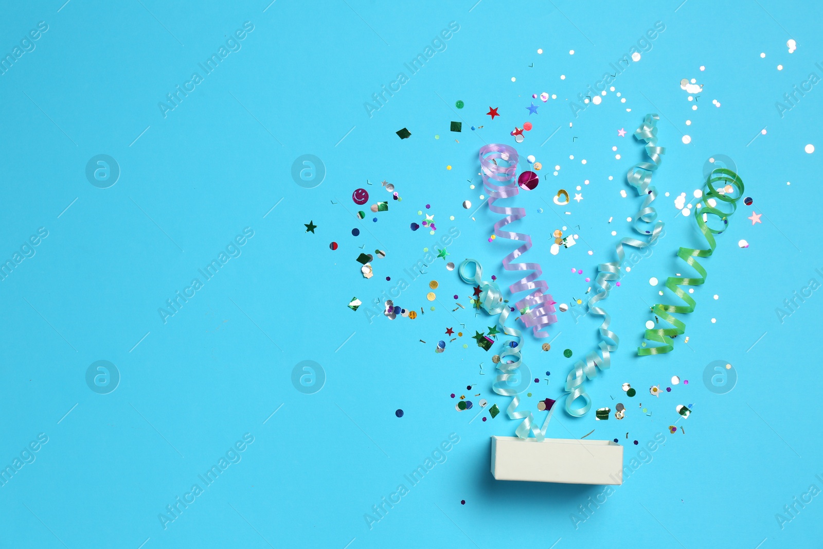 Photo of Shiny confetti and party streamers flying out of little box on light blue background, flat lay. Space for text