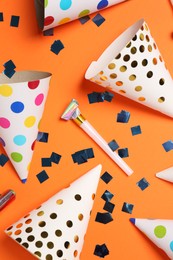 Shiny confetti, blowers and party hats on orange background, flat lay