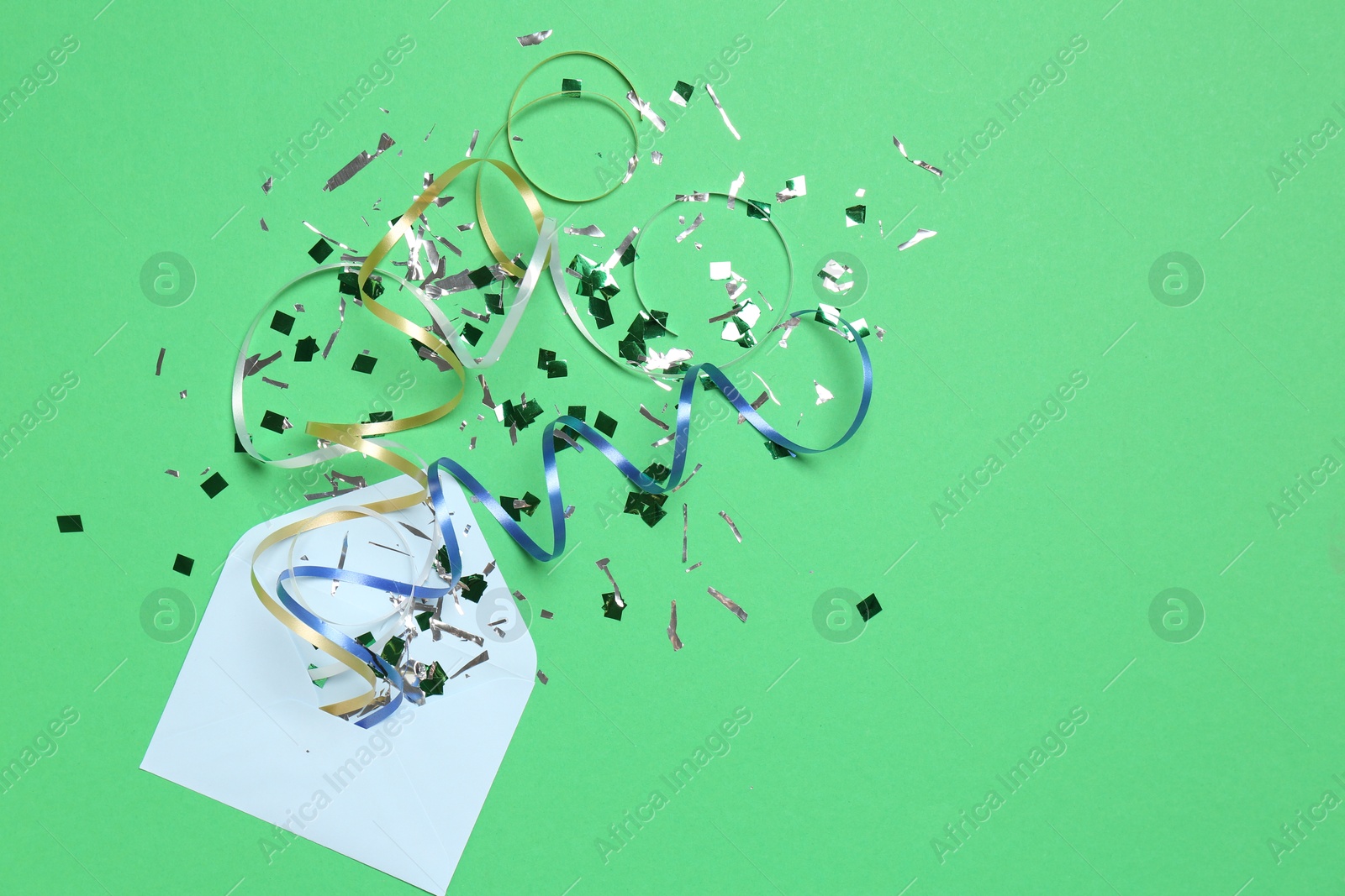 Photo of Shiny confetti and party streamers flying out of envelope on green background, flat lay. Space for text