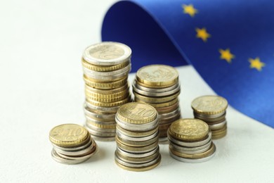 Photo of Stacks of euro coins and European Union flag on white textured table, closeup