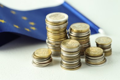 Photo of Stacks of euro coins and European Union flag on white textured table, closeup