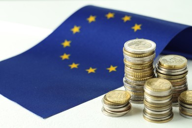 Photo of Stacks of euro coins and European Union flag on white textured table, closeup