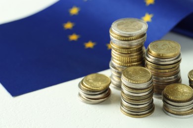 Photo of Stacks of euro coins and European Union flag on white textured table, closeup