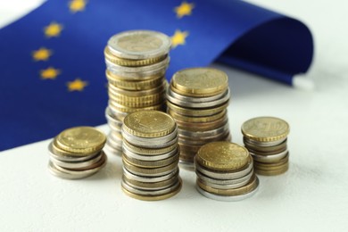 Photo of Stacks of euro coins and European Union flag on white textured table, closeup