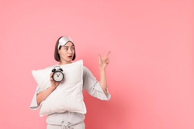 Photo of Overslept young woman with sleep mask, pillow and alarm clock pointing at something on pink background. Space for text