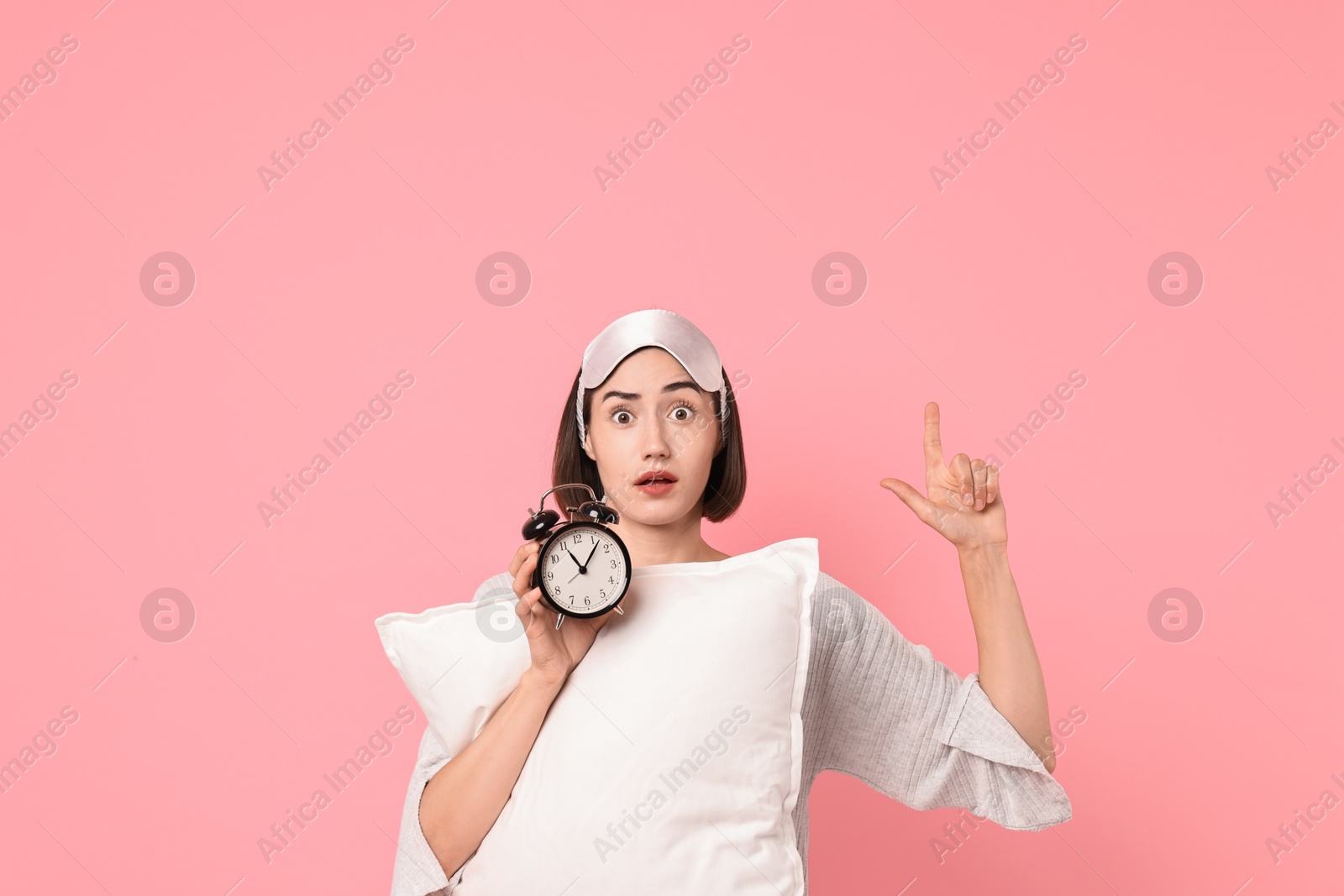 Photo of Overslept young woman with sleep mask, pillow and alarm clock pointing at something on pink background. Space for text