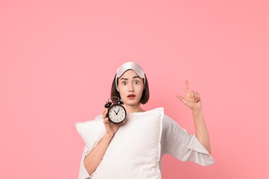 Photo of Overslept young woman with sleep mask, pillow and alarm clock pointing at something on pink background. Space for text