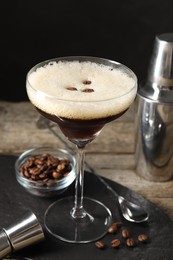 Photo of Glass of delicious espresso martini, jigger and coffee beans on wooden table against black background, closeup