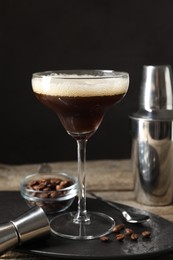 Photo of Glass of delicious espresso martini, jigger and coffee beans on wooden table against black background, closeup