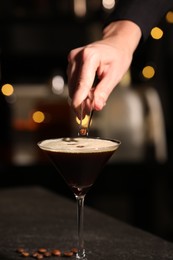 Bartender putting coffee bean into glass with delicious espresso martini at dark table against blurred lights, closeup